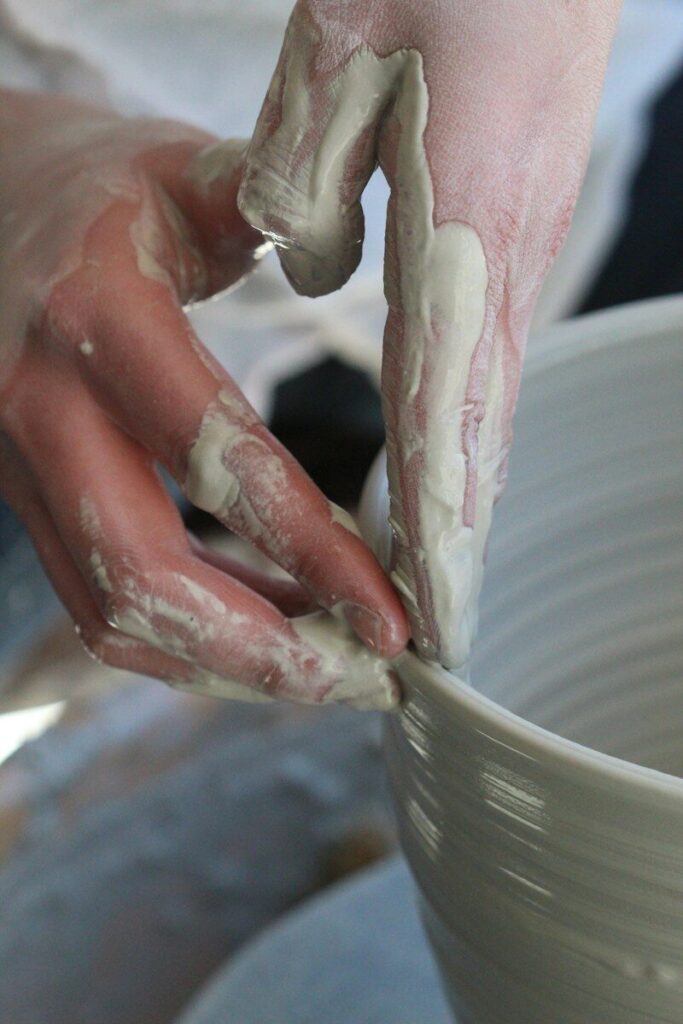 Hands covered in wet grey clay shaping the rim of a ceramic bowl